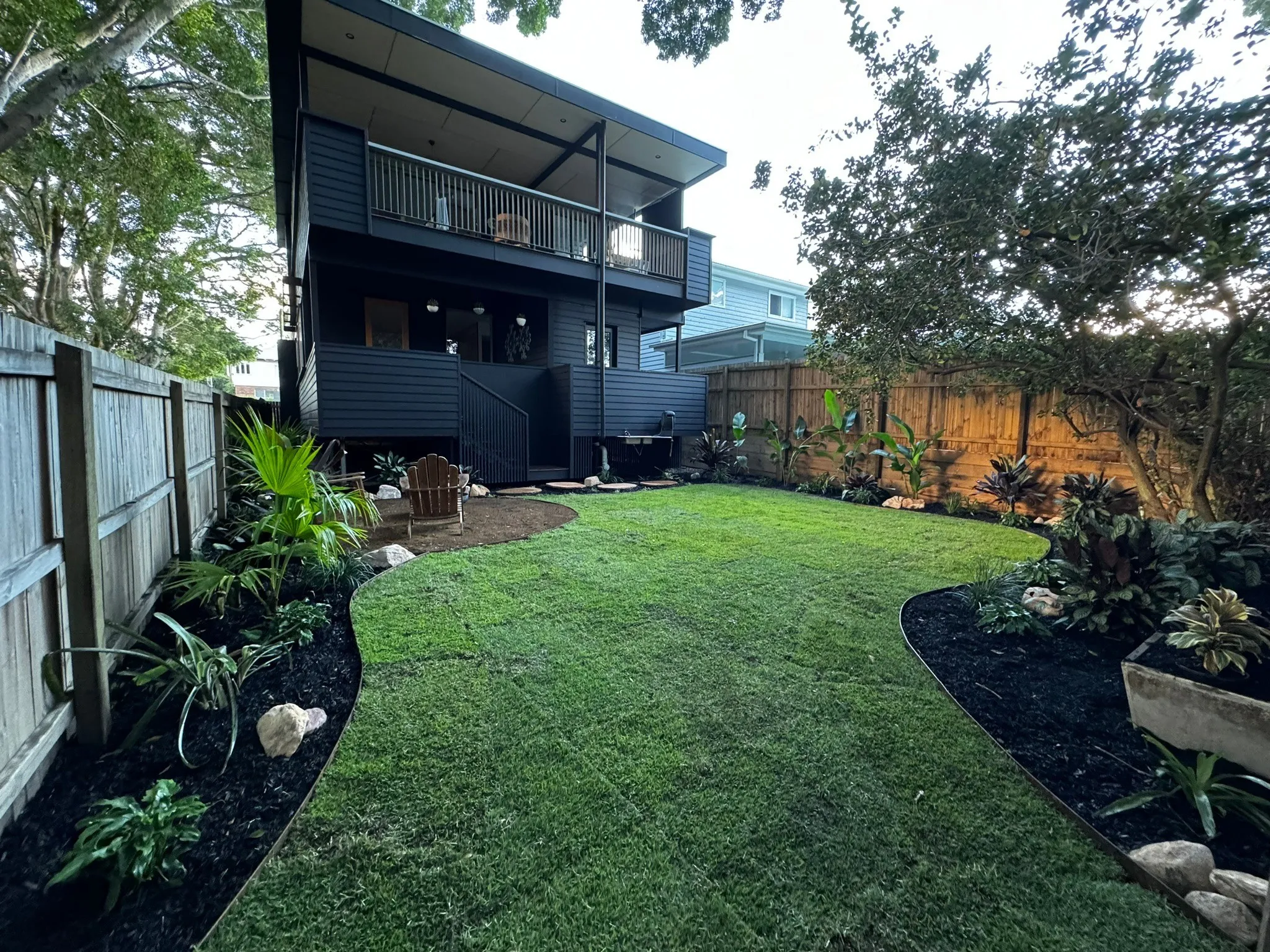 Backyard with freshly laid lawn curving between mulched tropical garden beds behind a dark two-storey home